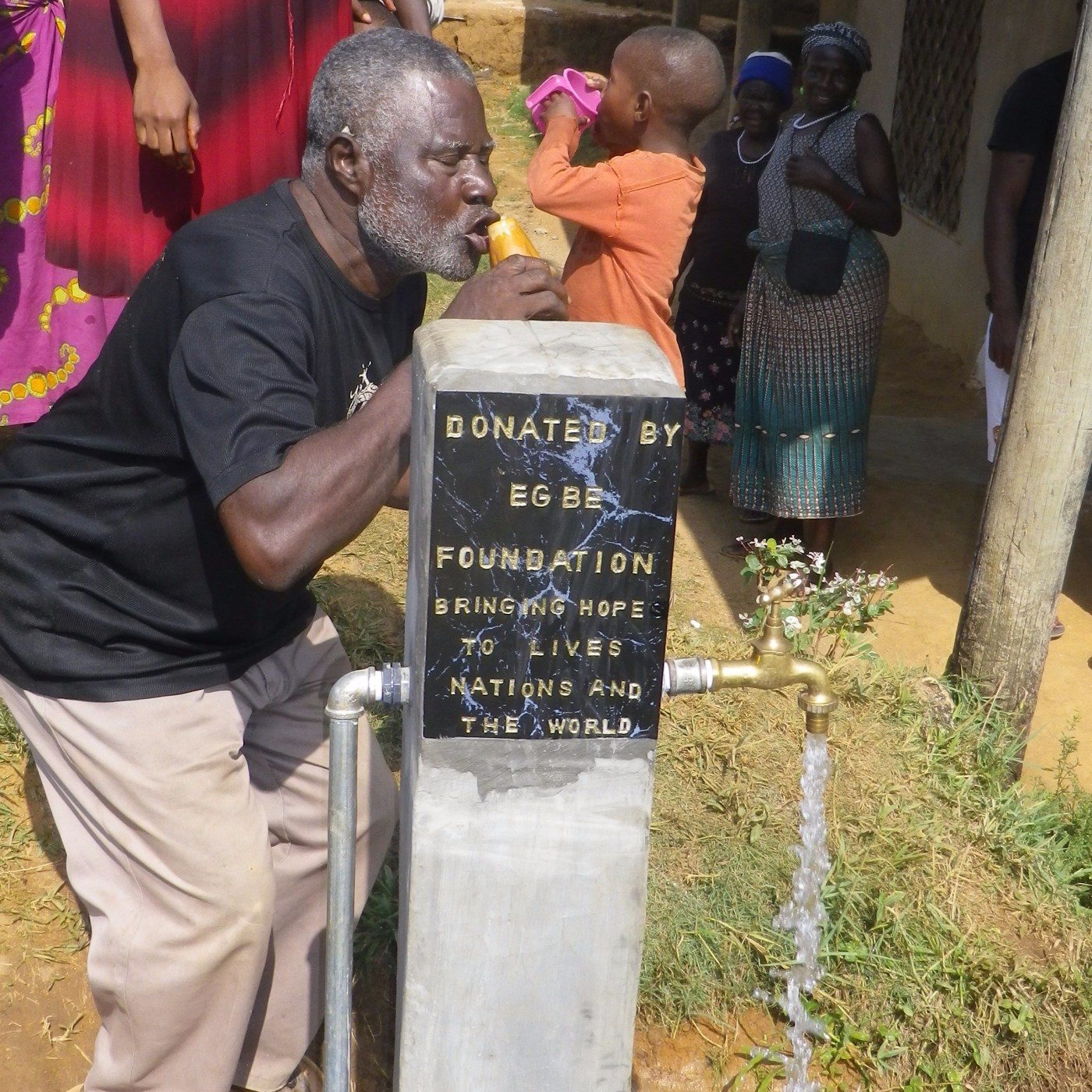 An elder drinks from a new public tap with a plaque reading Donated by Egbe Foundation